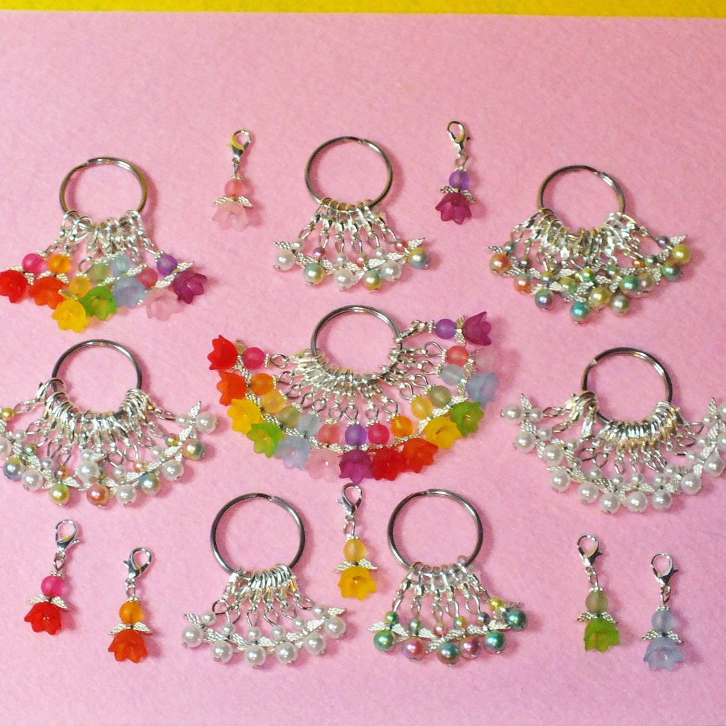 An image showing a range of handmade stitch markers. Each set comprises a keyring, on which hangs between 6-14 angels on clasps. There are a range of colours, white and rainbow mixes.
